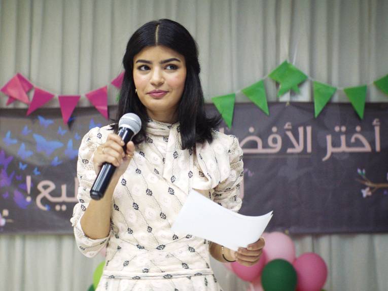 Im Bild zu sehen ist ein junge Frau mit Mikrofon und Notizen für eine Rede. Im Hintergrund deutet sich eine Partykulisse an und ein Transparent mit arabischer Schrift.