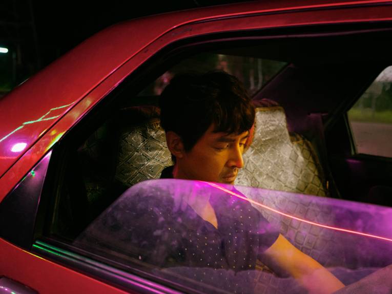 Im Bild zu sehen ist ein Mann in einem Auto, dessen Scheibe von pinkem Neonlicht angestrahlt wird.