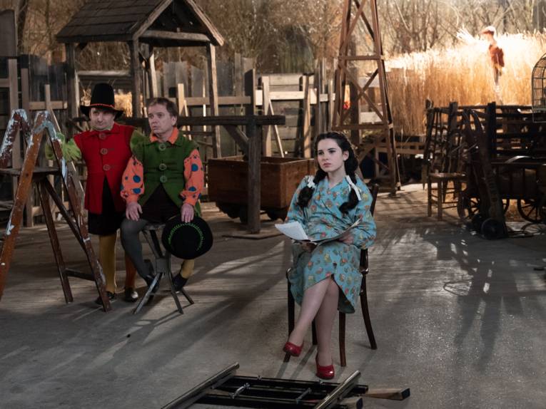 Im Bild zu sehen ist ein Filmset mit einigen kostümierten Schauspielern, darunter eine junge Frau im blauen Kleid mit roten Schuhen.