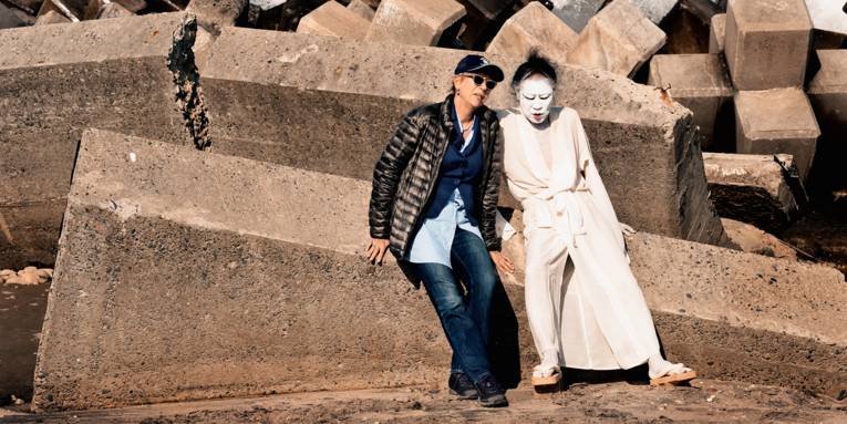 Regisseurin Doris Dörrie sitzt mit einer weiß gekleideten und im Gesicht weiß geschminkten Person auf einem großen Stein.