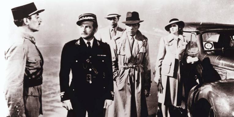 Im Bild zu sehen sind in Schwarz-Weiß einige Personen neben einem Auto auf einem Flugplatz, darunter Humphrey Bogart und Ingrid Bergman sowie zwei Uniformierte.