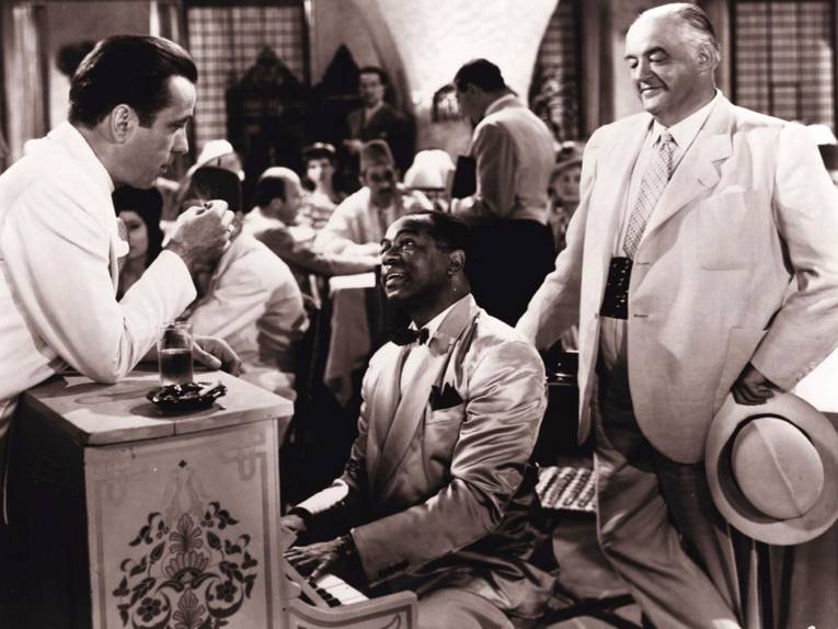 Im Bild zu sehen ist in Schwarz-Weiß ein exotisches Nachtlokal. In der Mitte ein dunkelhäutiger Pianist, neben ihm zwei weiße Männer in weißen Anzügen, einer von ihnen Humphrey Bogart, der sich auf das Klavier lehnt.