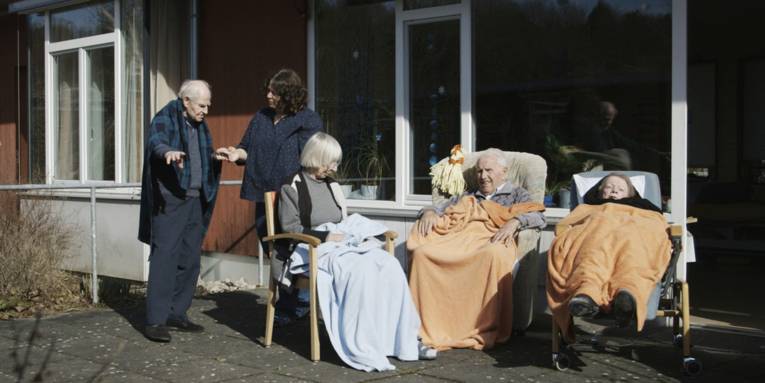 Im Bild zu sehen ist eine Gruppe Senioren, die auf einer Terrasse sind, einige sitzen unter Decken eingekuschelt in Stühlen. Vermutlich handelt es sich um eine Seniorenheim, eine Pflegerin steht bei einem älteren Mann.