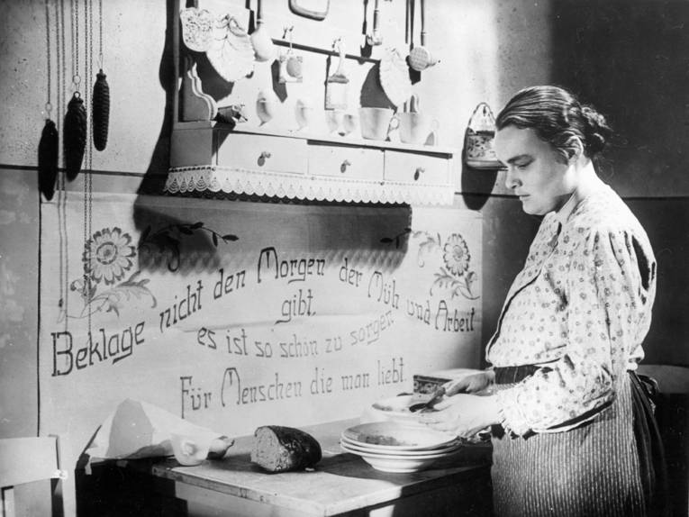 Mutter Bönike in der Küche. An der Wand vor ihr hängt der Spruch: Beklage nicht den Morgen, der Müh und Arbeit gibt, es ist so schön zu sorgen für Menschen, die man liebt