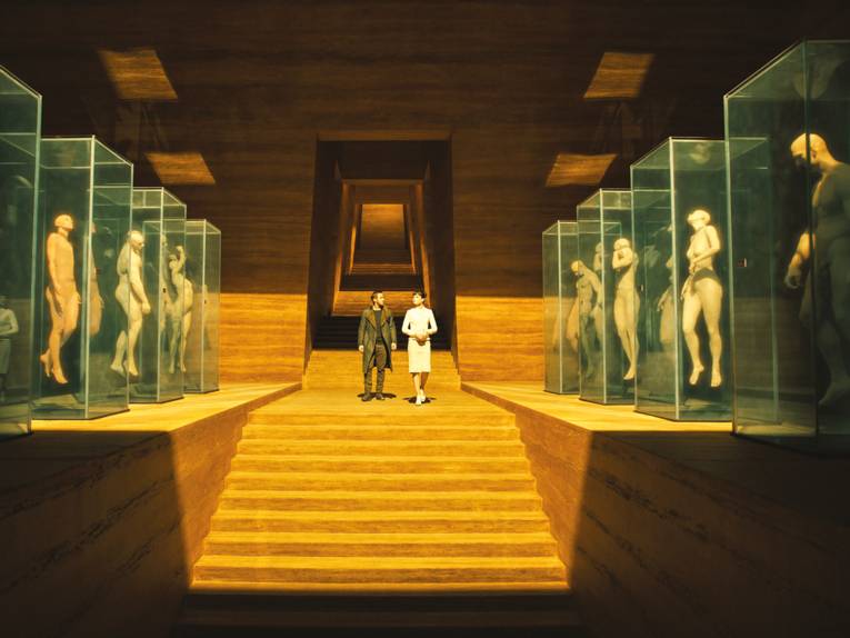 Ein großer Raum mit einer Treppe in der Mitte. Oben auf der Treppe stehen eine Frau und ein Mann. Rechts und links der Treppe stehen Glaskästen mit menschlichen Figuren.