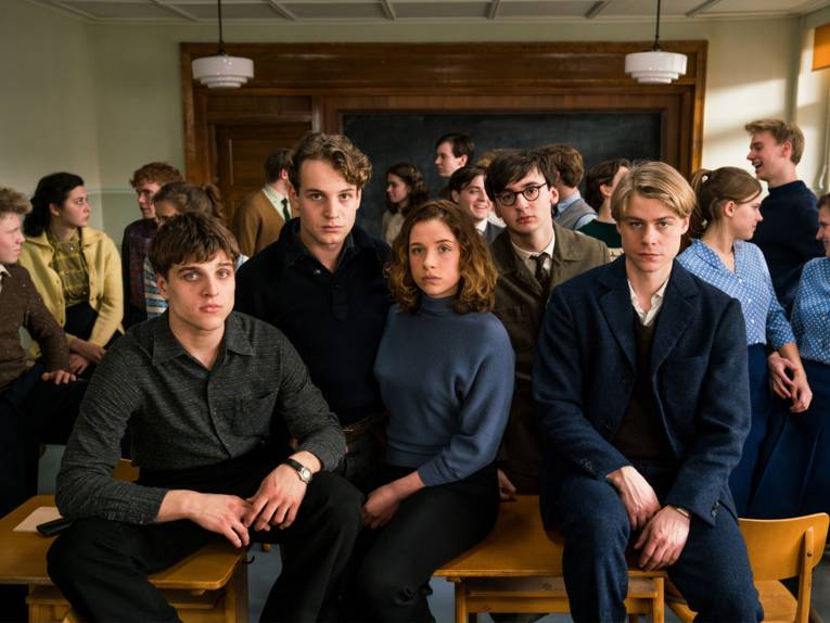 Fünf Schüler sitzen in einem Klassenraum und schauen in die Kamera. Hinter ihnen sind ihre Mitschüler am reden.