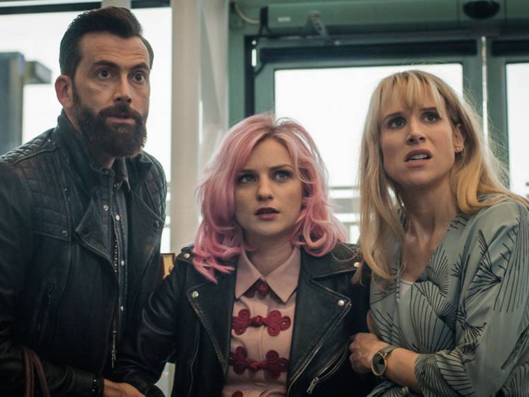 Im Bild zu sehen sind drei Personen, die etwas für den Betrachter nicht sichtbares gebannt anblicken. Rechts ist ein Mann mit Bart und Lederjacke, in der Mitte eine Frau mit rosa-gefärbter Frisur und links eine blonde Frau, die beunruhigt wirkt.