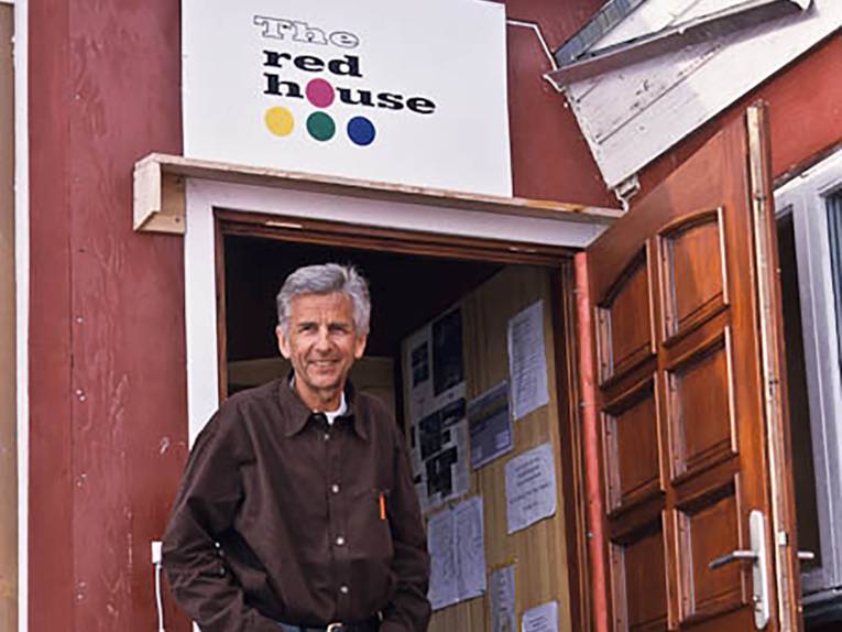 Robert Peroni, ein schlanker Mann mit grauem Haar, steht lächelnd in einer offenen Eingangstür. Über der Tür hängt ein Schild, auf dem The red house steht.