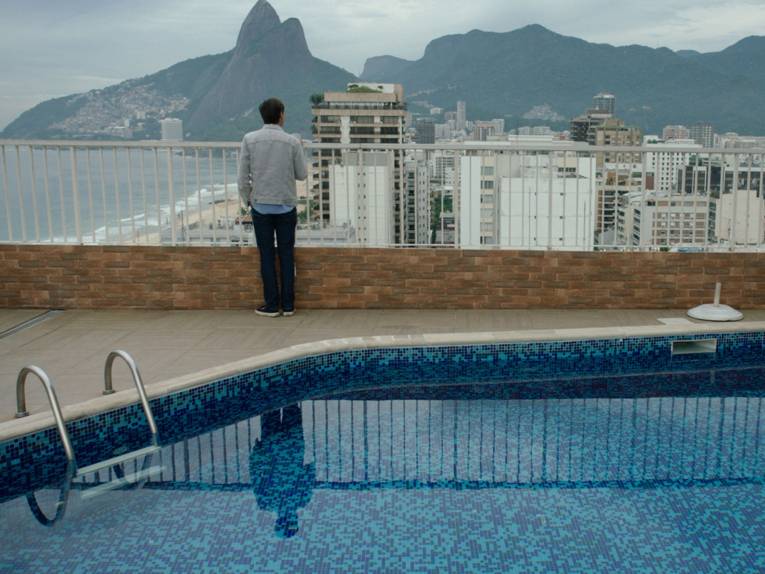 Mann steht hinter einem Pool an einem Geländer und schaut von oben auf eine Stadt.