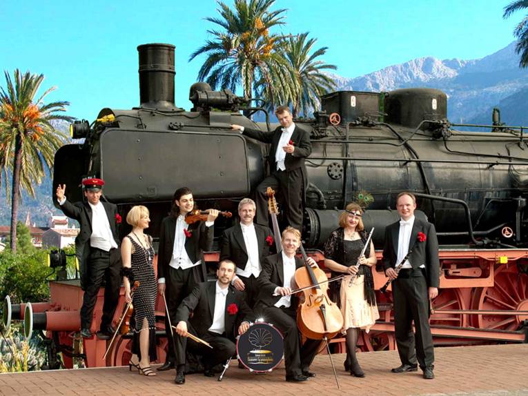 Das Salonorchester Hannover Grammophons, zwei Frauen und sieben Männer, stehen mit ihren Instrumenten vor einer alten Dampflokomotive auf Mallorca - im Hintergrund sind Palmen und ein Gebirgszug zu sehen