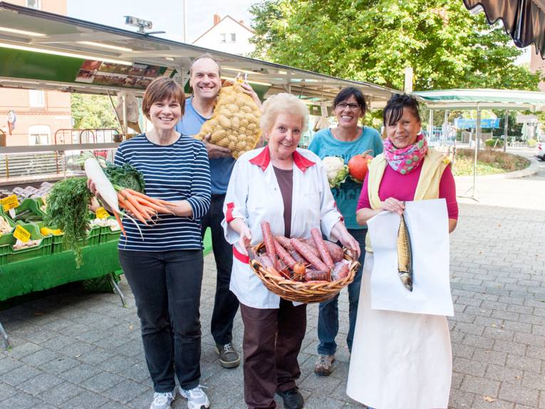 Ein Markthändler und vier Markhändlerinnen präsentieren markttypische Erzeugnisse: Kartoffeln, Wurst, Kürbis, Blumenkohl, Möhren, Rettich und Fisch