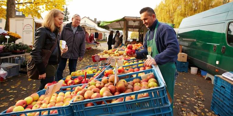 Kundin und ein Interessent vor einem Markstand mit Äpfeln; der Obstverkäufer hält vier Äpfel in einer Hand.