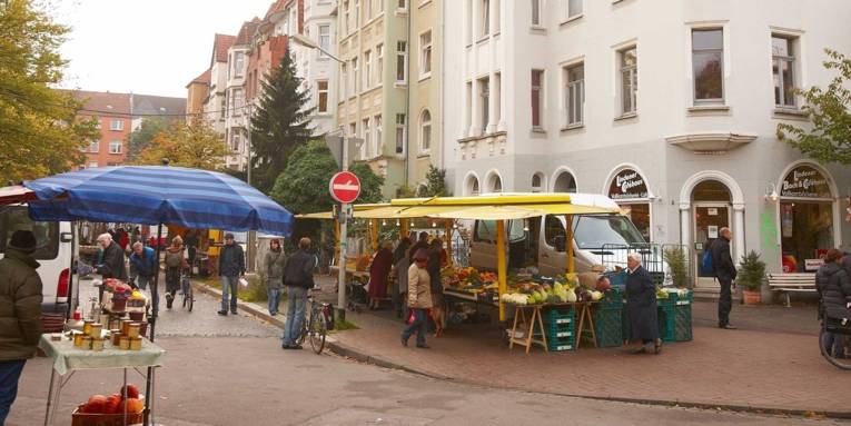 Vorne links ein kleiner Stand mit Honig und Kürbissen, gegenüber ein Obst- und Gemüsestand, an dem einige Kundinnen und Kunden stehen. Im Hintergrund Jugendstil-Häuser.