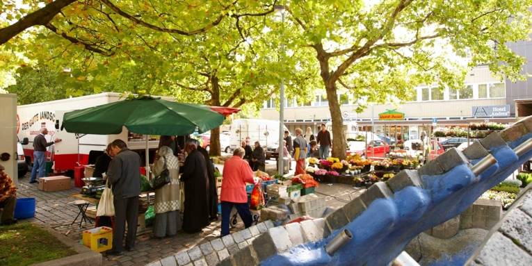 Marktbesucherinnen an verschiedenen Wochenmarktständen; im Vordergrund der Stöckener Brunnen, im Hintergrund Platanen und Geschäftshäuser