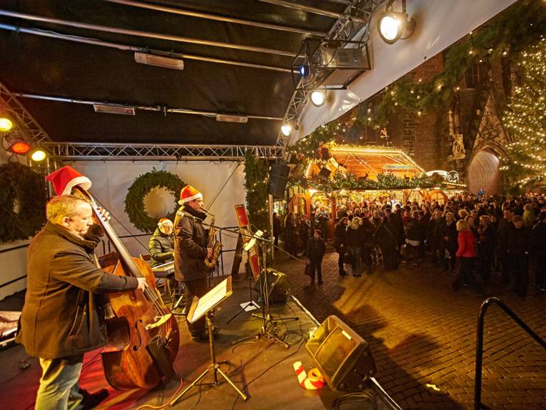 Drei Musiker (Bass, Saxofon und Keyboard) spielen mit Weihnachtsmützen vor Publikum auf einer Bühne an der Marktkirche