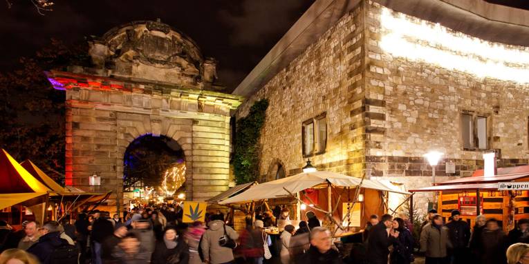 In der vorweihnachtlichen Dunkelheit sorgen der angestrahlte Torbogen und die Mauer des Historischen Museums für eine stimmungsvolle Kulisse der rundherum aufgebauten Marktstände, die von zahlreichen Besucherinnen und Besuchern frequentiert sind.