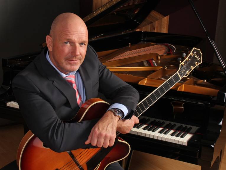 Ein Mann in einem Anzug sitzt auf einem Klavierschemel an einem Klavier und hält dabei eine Gitarre im Arm.