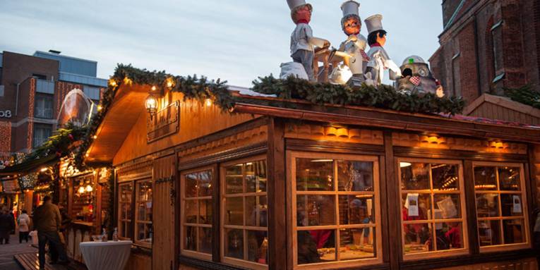 Auf dem Dach einer weihnachtlich geschmückten Holzhütte stehen Puppen in Bäckerkleidung; innen sind Kinder an einem großen Tisch damit beschäftigt, Weihnachtskekse auszustechen.