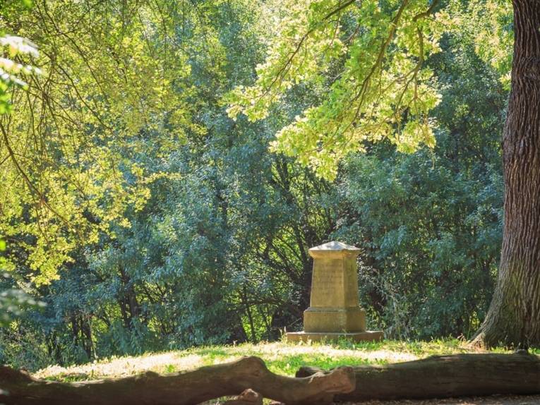 Auf einer Rasenfläche im Hinüberschen Landschaftspark steht eine Stele umrahmt von alten Bäumen