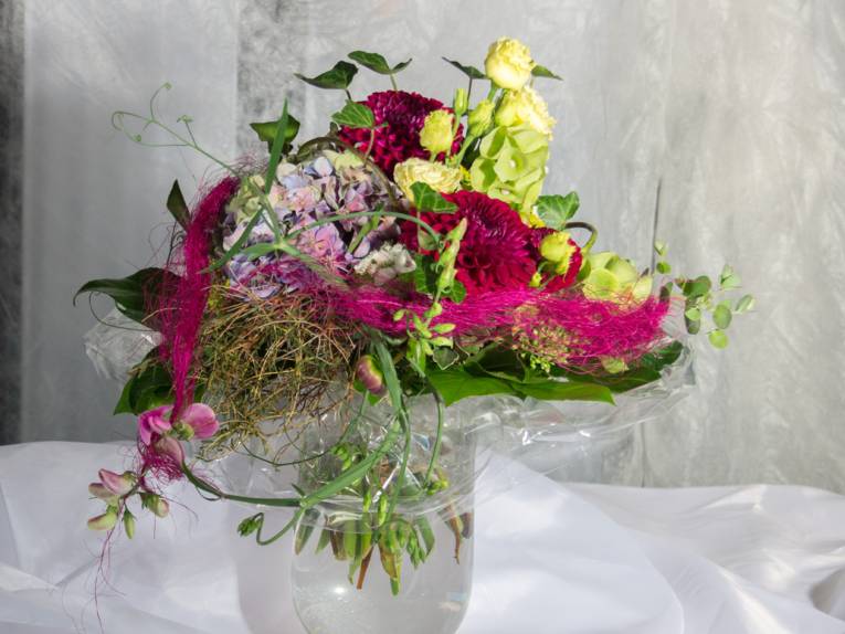 Ein Blumenstrauß mit Hortensienblüte, Rosen, Dahlien, Wicke, Efeu und Zierelementen in einer Glasvase