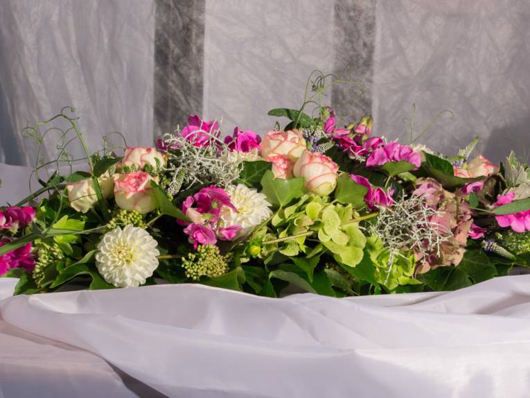 Ein Tischgesteck mit Dahlien, Rosen, Wicken, Hortensienblüte, Efeu auf weißem fließenden Tuch