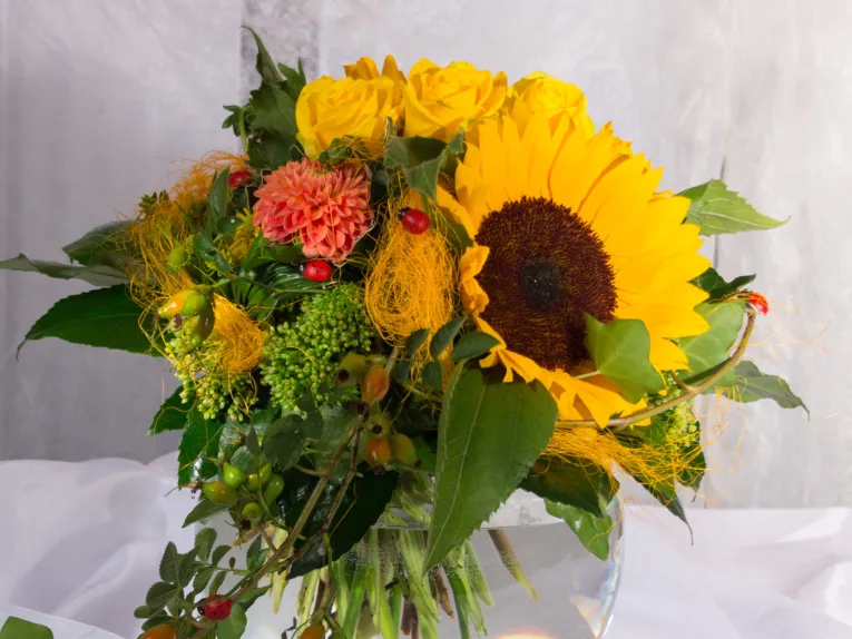 Ein Blumenstrauß in Gelbtönen mit roten Akzenten: Sonnenblume, Dahlie, Rosen, Hagebutten und Sedum mit Blattgrün werden von roten Marienkäfern geziert.