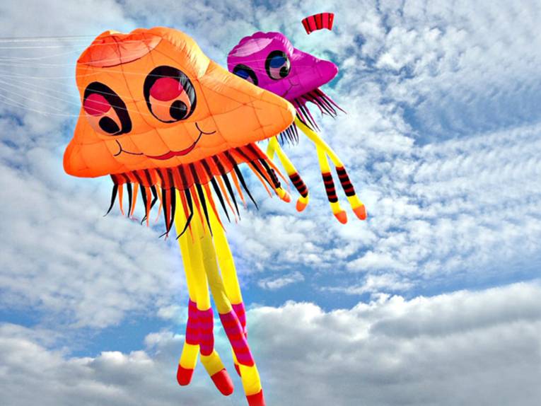 Zwei bunte Drachen, die wie lächelnde Kraken aussehen, fliegen vor blauem Himmel mit Schäfchenwolken