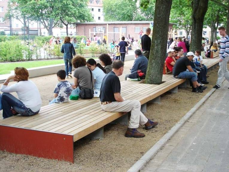 Kinder und Erwachsene sitzen auf Holzbänken, die an einen Spielplatz angrenzen