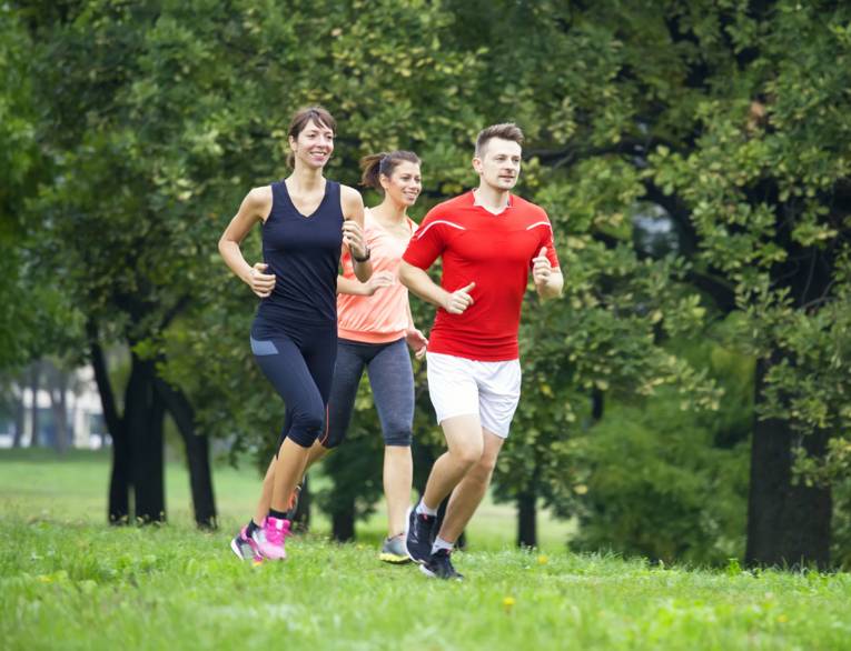Zwei junge Frauen und ein junger Mann joggen in Sportkleidung durch eine Grünanlage