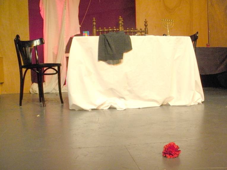 Auf einer Bühne steht ein Tisch mit zwei Stühlen und auf dem Boden liegt eine rote Nelke
