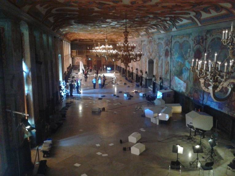 Das barocke Galeriegebäude vom Balkon ausgesehen; Besucher laufen durch die Videoskulptur von caprificus.org. Weiße Blätter liegen auf dem Boden.