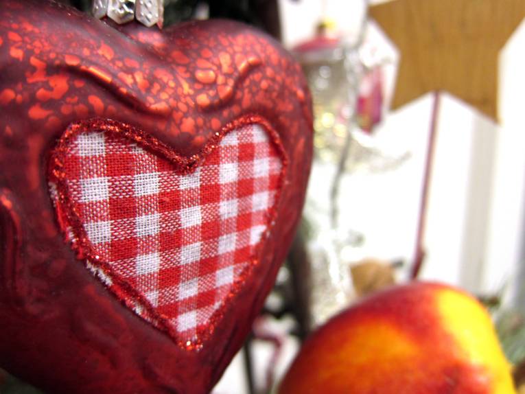 Eine herzförmige Weihnachtskugel an einem kleinen Weihnachtsbaum, im Hintergrund sind ein sternförmiger Anhänger sowie ein künstlicher Apfel zu erkennen