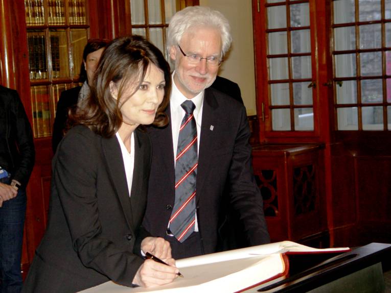 Iris Berben und Bernd Strauch stehen an einem Pult, auf dem das Goldene Buch der Stadt liegt. Berben trägt sich ein und schaut gleichzeitig in Richtung der Fotokameras.