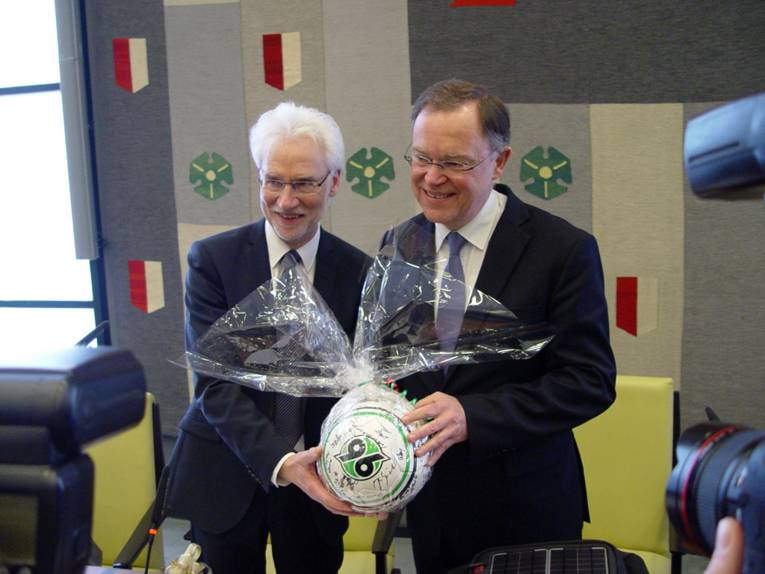 Bernd Strauch und Stephan Weil halten einen signierten Fußball in der Hand, drumherum Kamera-Objektive