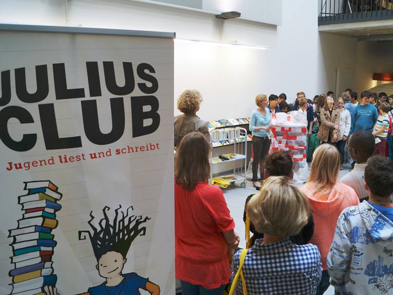 Eine Gruppe Jugendlicher steht im Halbkreis um eine etwas ältere Frau, die gerade spricht, links im Bild ist ein Aufsteller für den JULIUS-CLUB