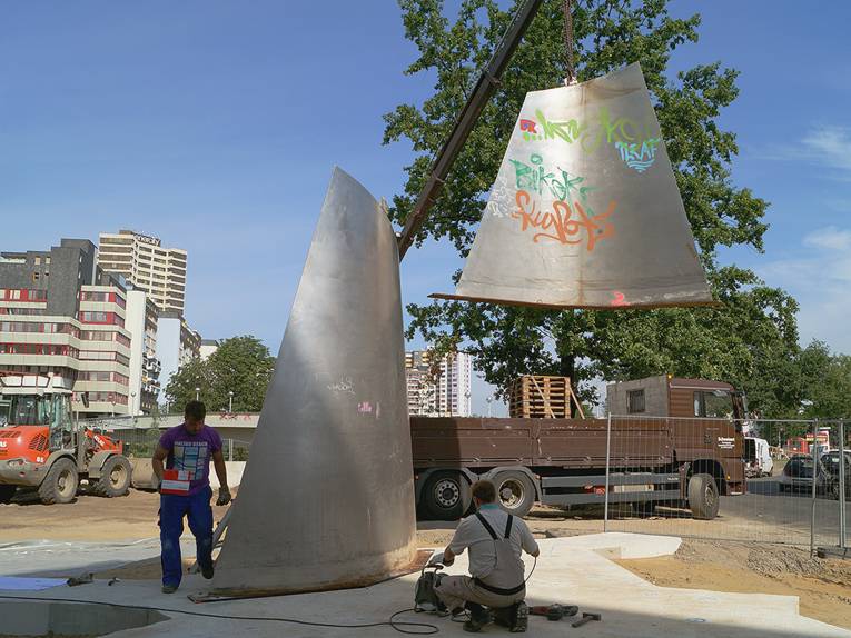 Ein Teil einer Metall-Skulptur wird mittels eines Krans auf ein Beton-Fundament gesetzt
