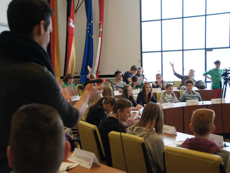Blick in die Ratssitzung des Planspiels "Pimp Your Town!", ein Schüler im Vordergrund spricht gerade, im Hintergrund gibt es mehrere Wortmeldungen