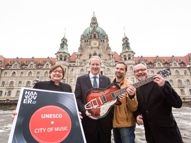 Marlis Drevermann, Stefan Schostok, Steffen König und Olaf Neumann mit einer Gitarre und einem Plakat "UNESCO City of Music" vor dem Neuen Rathaus