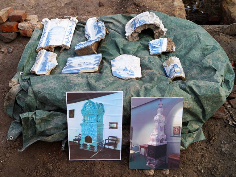 Reste eines Kachelofens: Blau-weiße Fliesen, davor zwei Fotos von intakten Öfen