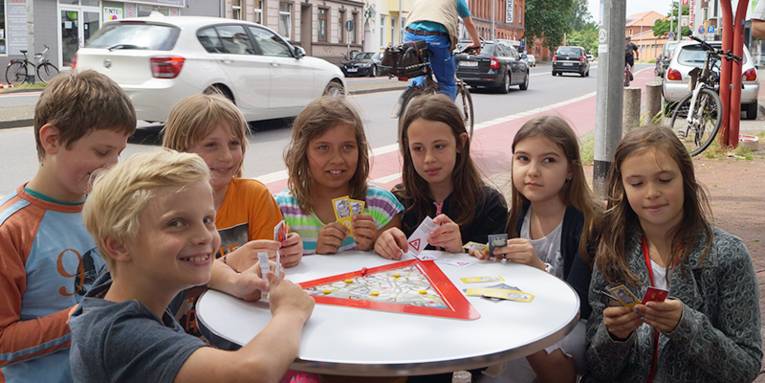 Schülerinnen und Schüler der 4. Klasse der Bonifatius-Grundschule spielen das neue Brettspiel "Verkehrsmonsterjagd" auf einem Tisch an einer befahrenen Straße