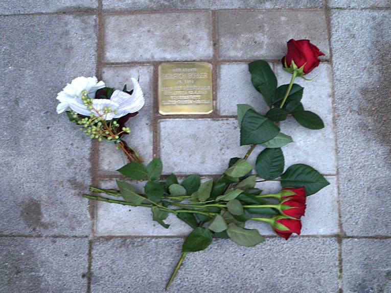 Der quadratische messingfarbene Stolperstein für Heinrich Börner, der ins Straßenpflaster eingelassen wurde, wird von roten Rosen umrahmt.