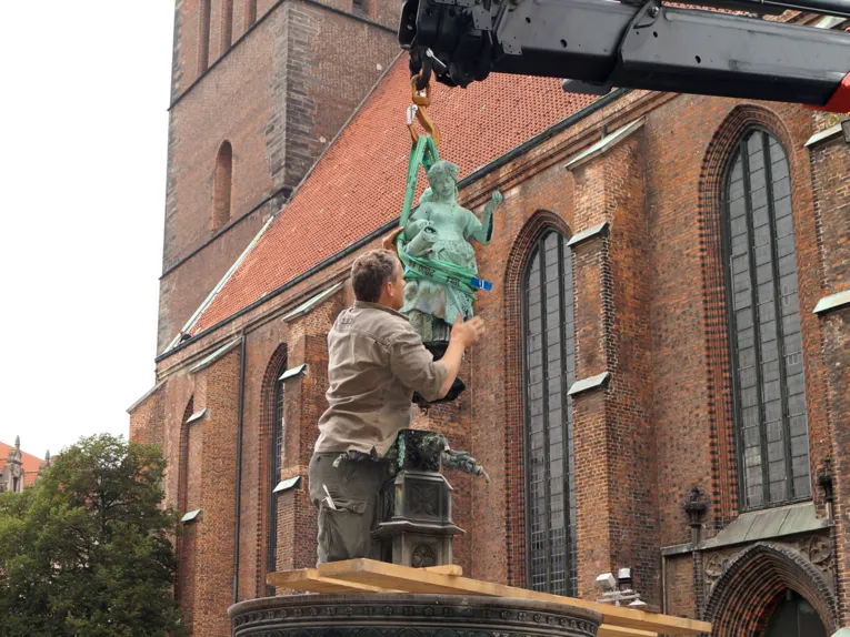 Ein Mitarbeiter der Stadt steht im Brunnen und achtet darauf, dass die an einem Kran schwebende Frauenfigur keinen Schaden nimmt