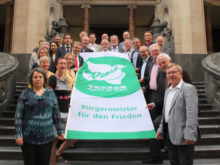 Knapp 30 Personen zeigen die Flagge der "Mayors for Peace"
