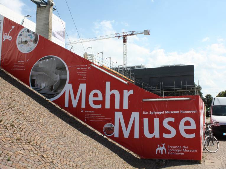 Der Bauzaun am Sprengel Museum mit Baukran und dem dunklen quaderförmigen Erweiterungsbau im Hintergrund