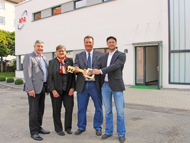 Eine Frau und drei Männer stehen vor einem Gebäude und halten symbolisch einen goldenen Schlüssel in die Kamera