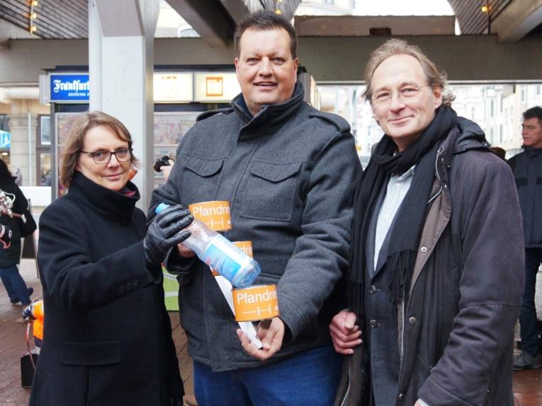 Eine Frau und zwei Männer, die einen Ring und eine Flasche zeigen. 