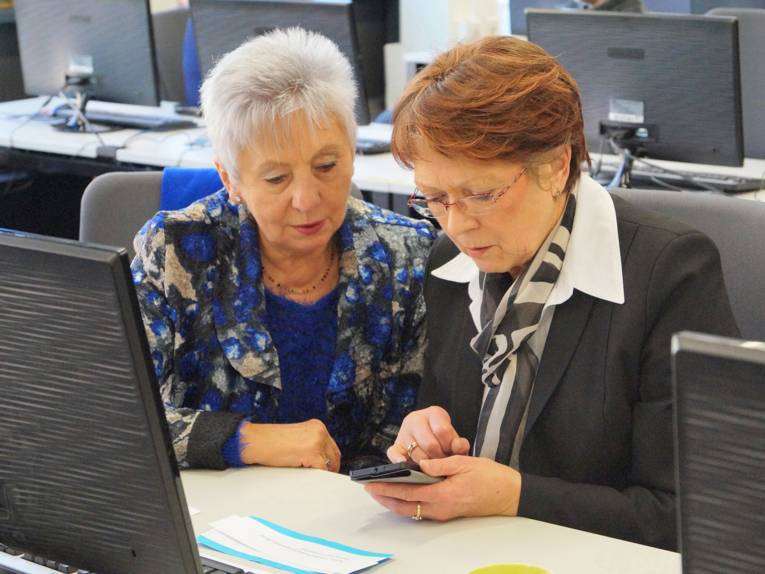 Zwei ältere Damen blicken auf ein Smartphone, während sie vor einem PC sitzen