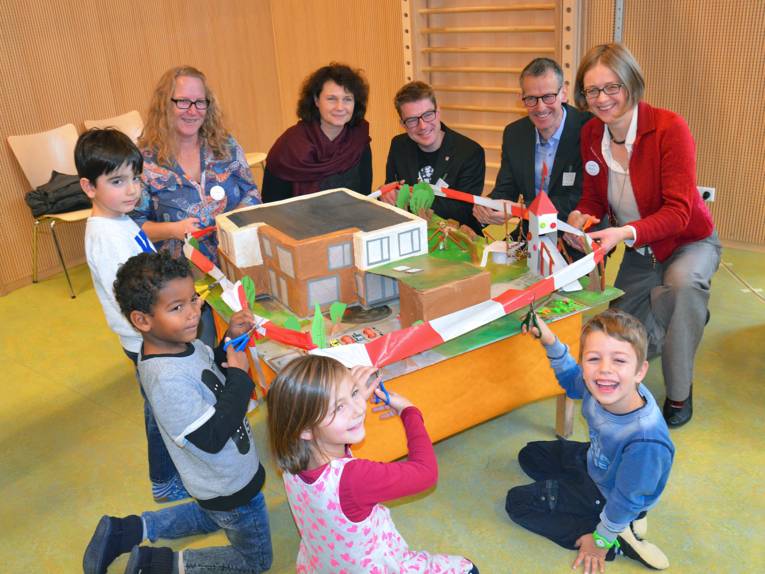 Mehrere Personen hocken um das Modell einer Kita und schneiden zur Einweihung rot-weißes Flatterband durch