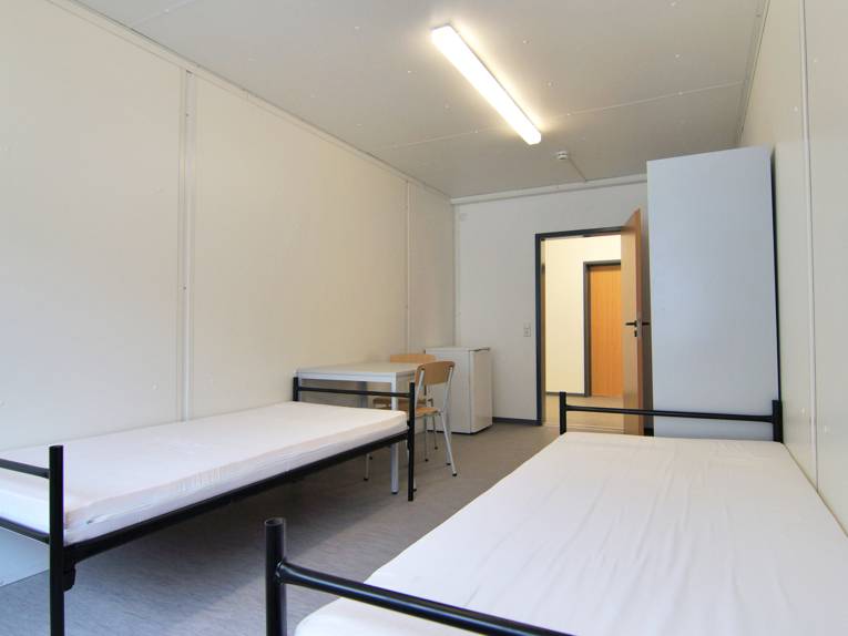 Zwei Betten, ein Tisch, zwei Stühle, die Seite eines Spindes und ein Kühlschrank in einem Zimmer der Flüchtlingsunterkunft Waterlooplatz
