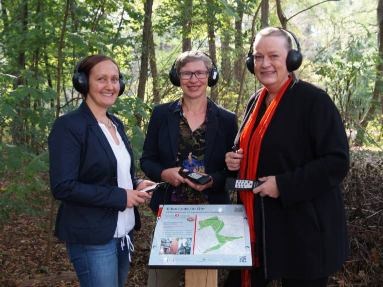 Drei lächelnde Frauen hinter einer Tafel im Wald. 